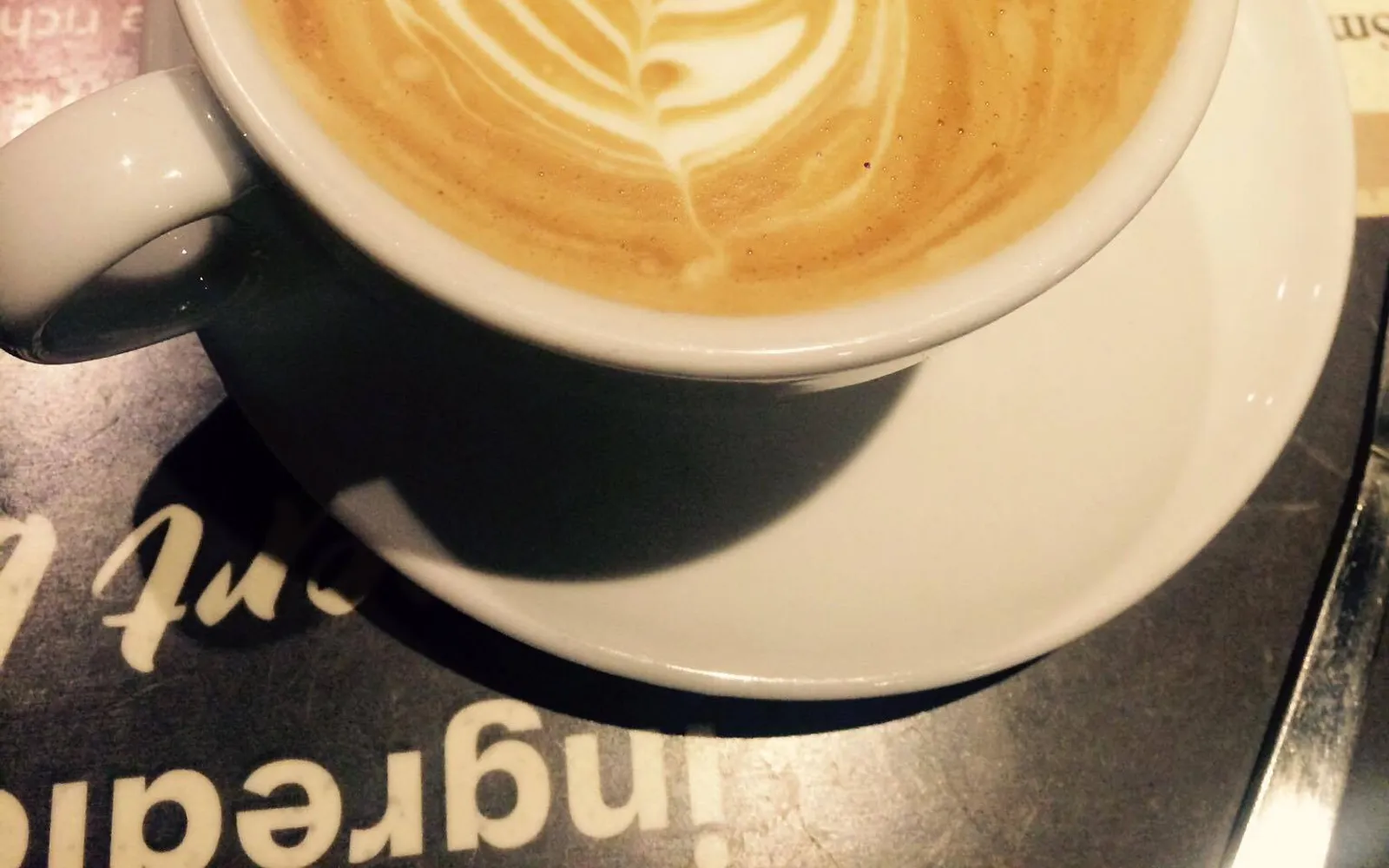 A flat white in a white ceramic cup with leaf latte art on a thin layer of micro-foam, served on a white plate with a small piece of chocolate