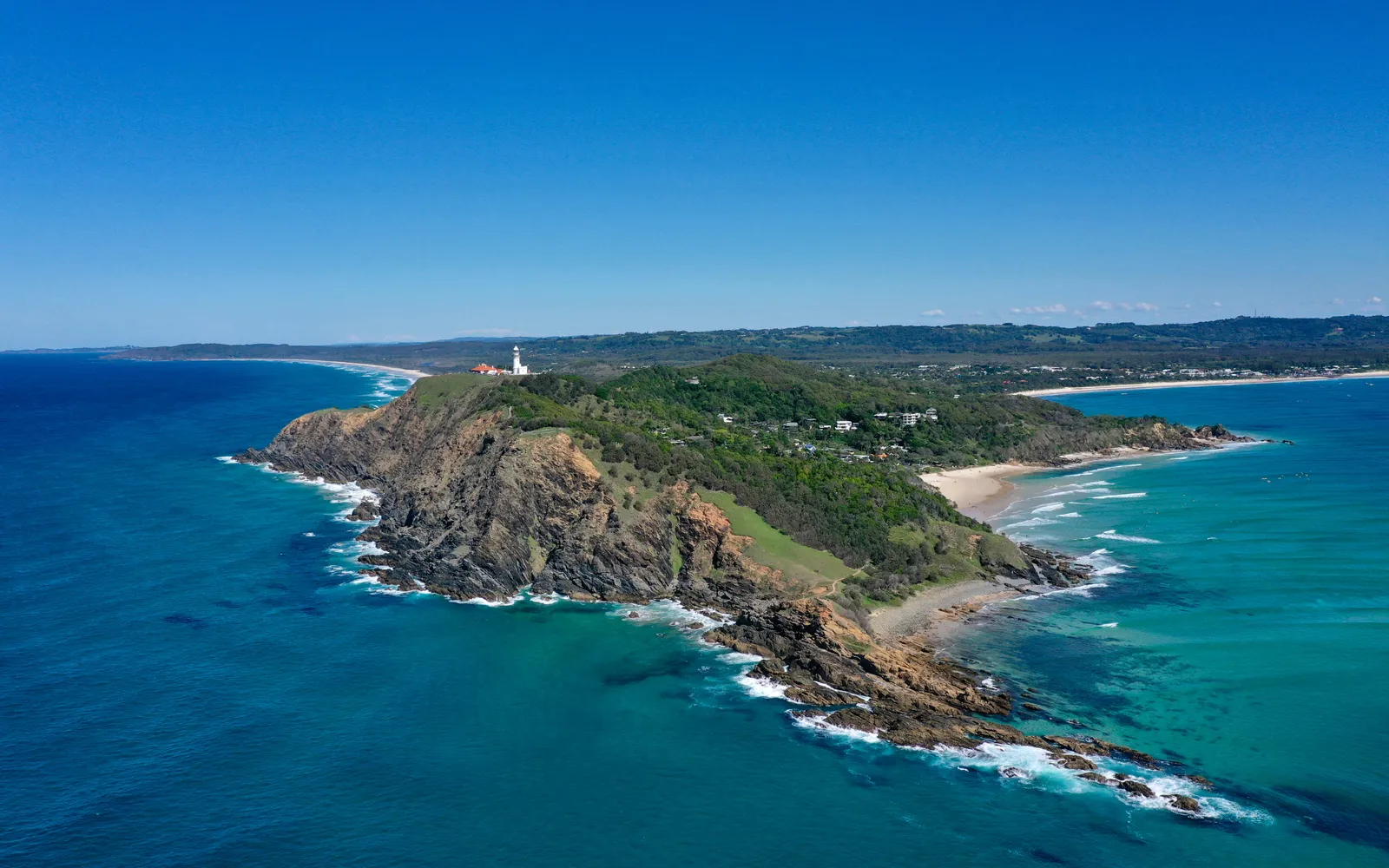 Byron Bay's Main Beach in the morning, a long stretch of white sand, swimmers in the water, surfers paddling their boards, a clear blue cloudless sky