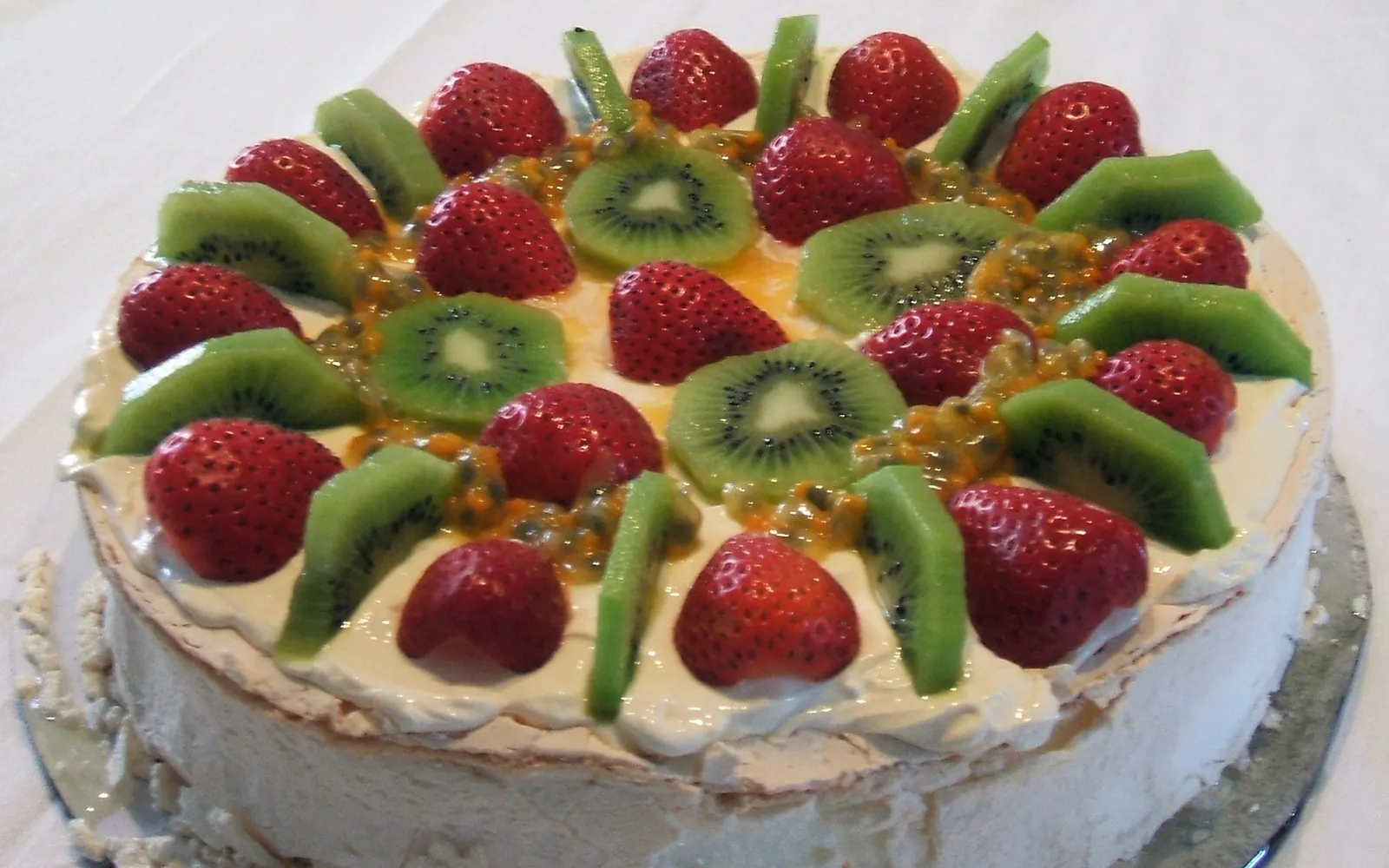A large pavlova with a crisp cream-white shell, topped with whipped cream and bright fresh fruit — strawberry, kiwi and passionfruit