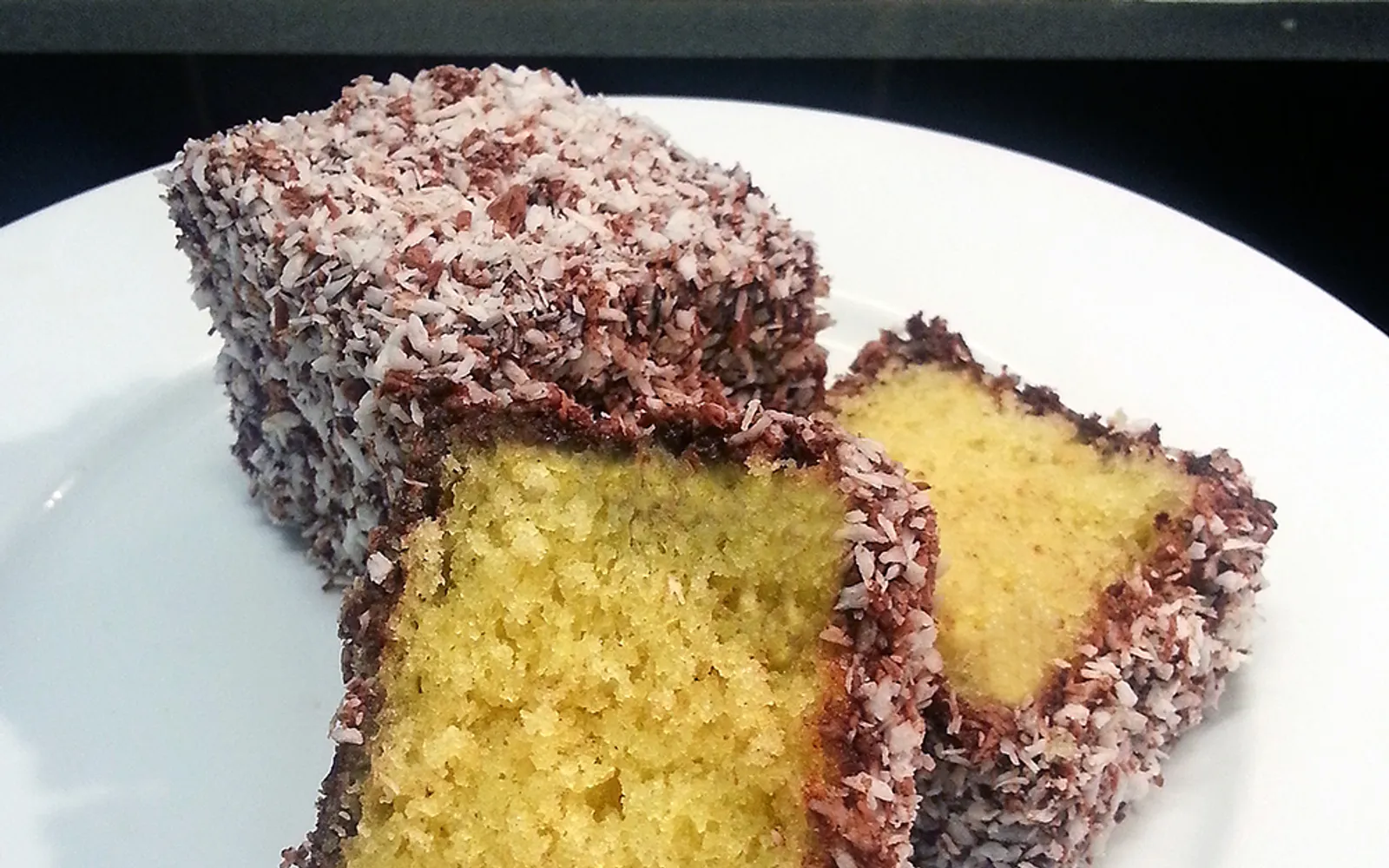 A square Australian lamington coated in chocolate and rolled in shredded coconut, cut in half to show the soft sponge inside