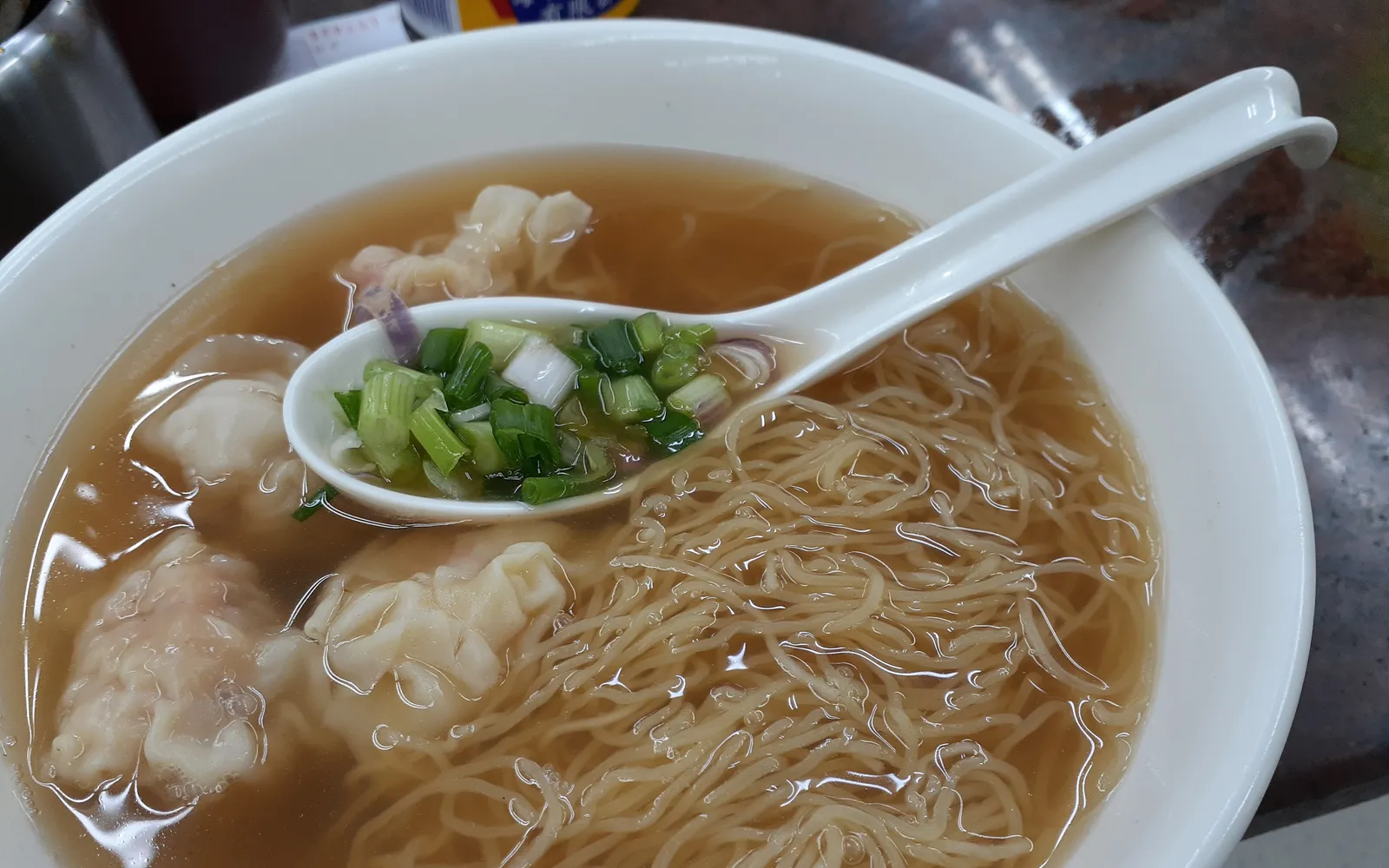 A bowl of Cantonese-style wonton noodles, thin translucent wontons in clear broth with soft yellow noodles