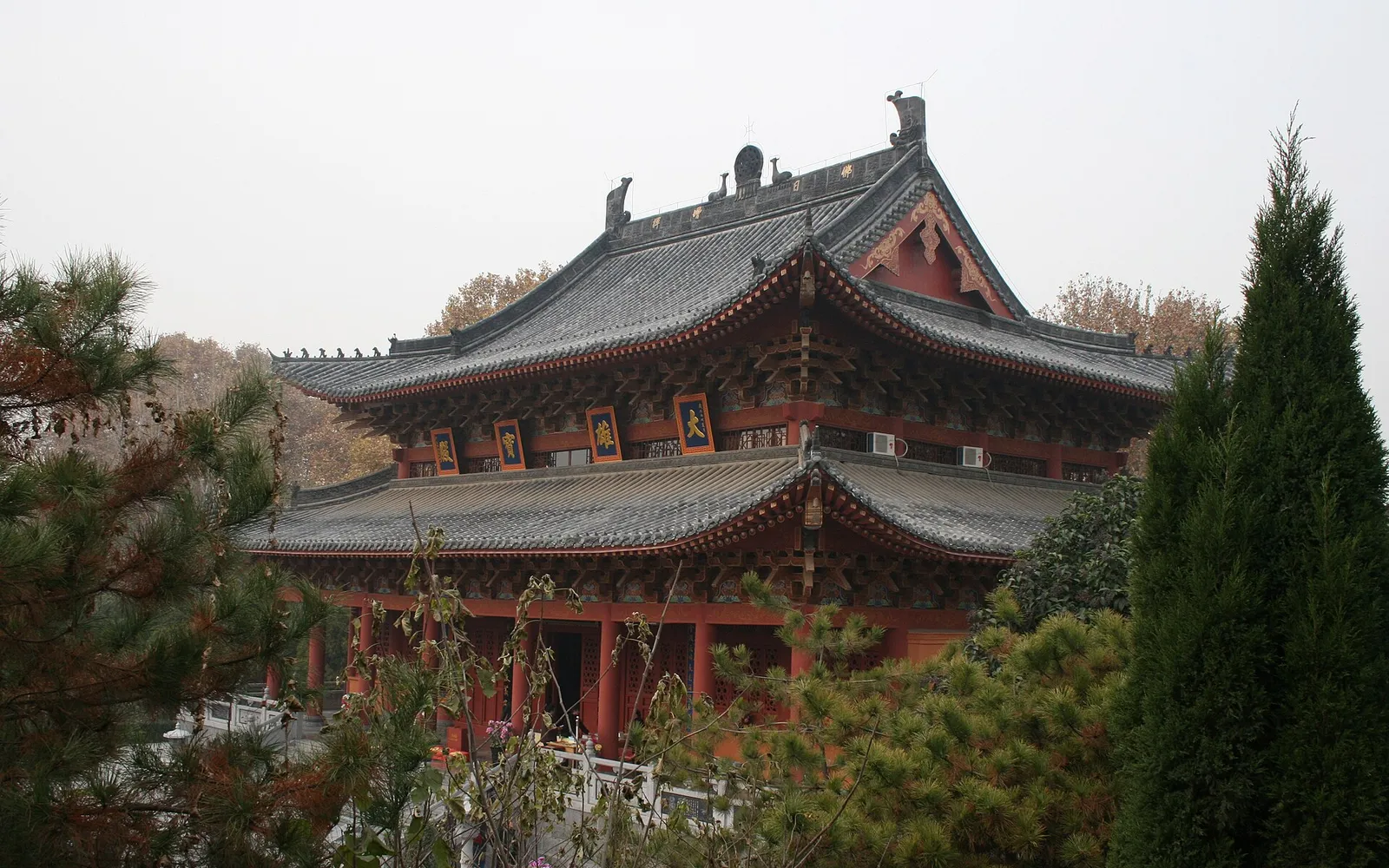 The deep-red entrance gate of the White Horse Temple decorated with carved dragons and phoenixes, with the temple's tall blue Qiyun Pagoda standing out behind it against a blue sky