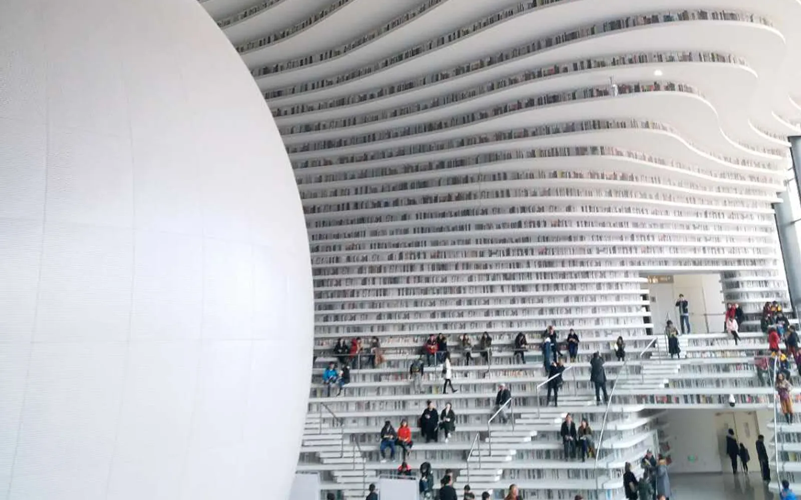 Interior of Binhai Library: white wave-like bookshelves curve from floor to ceiling across 5 levels, surrounding a silver spherical auditorium at the centre