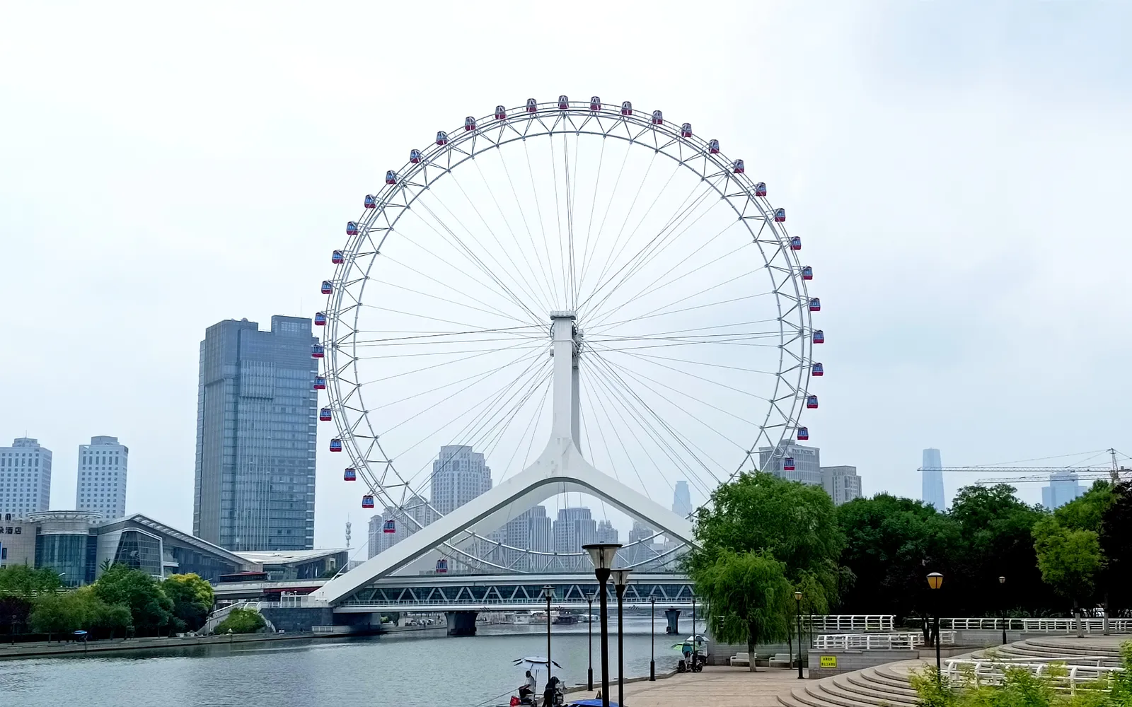 The Tianjin Eye ferris wheel sitting atop Yongle Bridge over the Hai River, white lights reflecting in the water at night with the city skyline behind