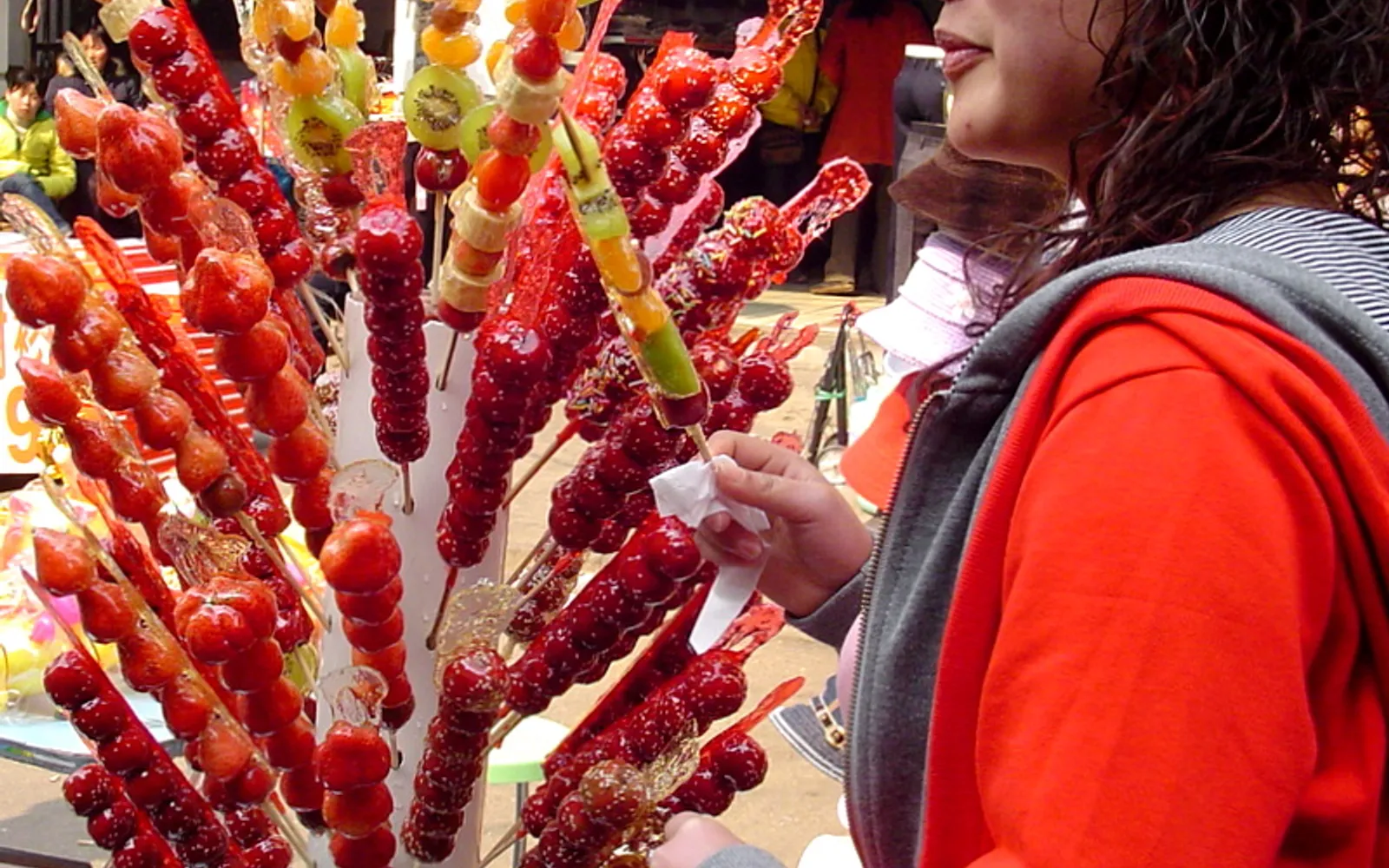 Skewers of candy-coated fruit — gleaming red sugar shells catch the light at a night-market stall, the coating as clear and bright as glass