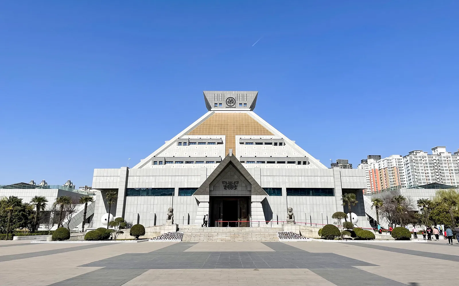 The Henan Museum's large pyramid-shaped building in golden brown stands prominently in a green park, its form inspired by ancient Chinese burial mounds