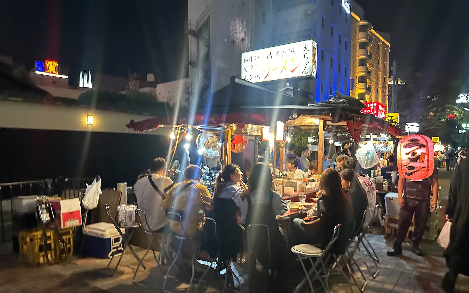 Brightly lit yatai stalls glowing in the Fukuoka night, with diners seated outside at the counter