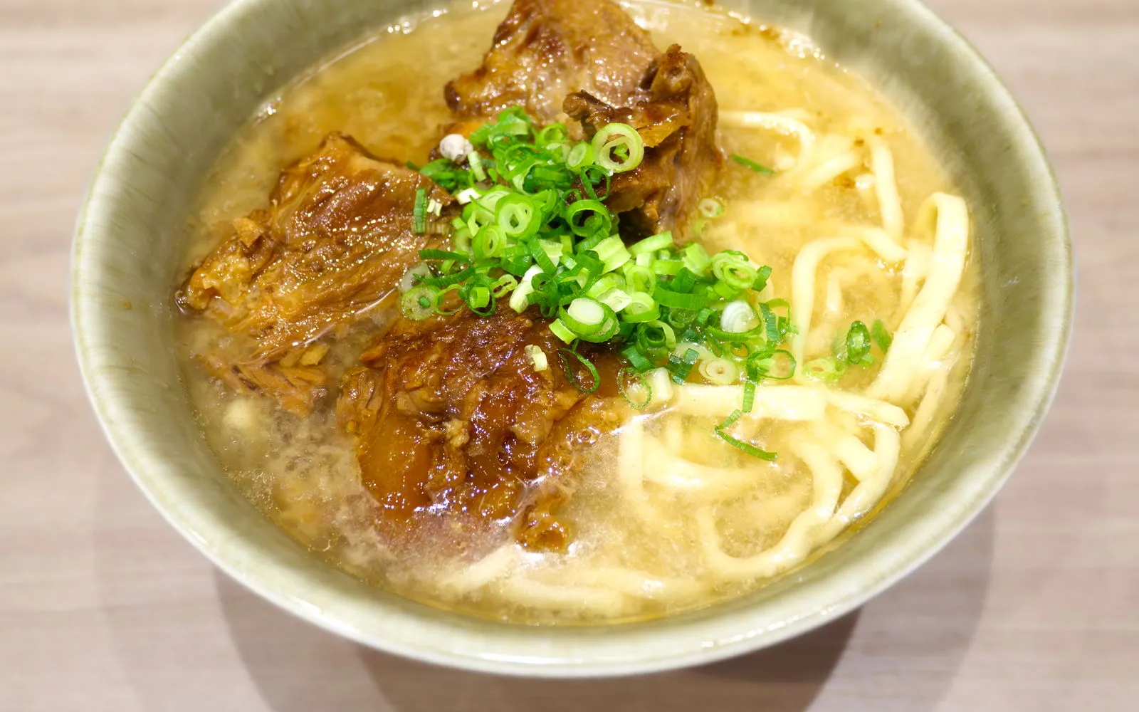 A bowl of Okinawa soba with golden clear broth, thick wheat noodles, braised pork belly, fish cake, and pickled ginger