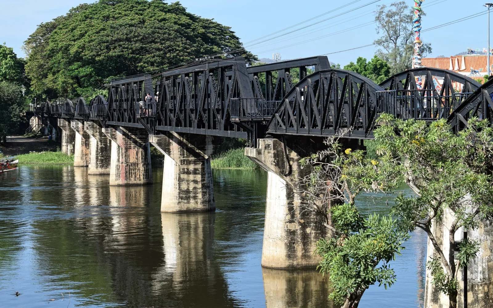 The Tham Krasae Railway Bridge curving along the cliff face above the Khwae Noi River, backed by green forest